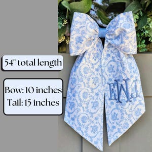 May include: A large decorative bow featuring a light blue and white floral pattern with a central blue knot. The fabric has a 54-inch total length, a 10-inch bow width, and 15-inch tails. A blue embroidered monogram is visible on the right tail.