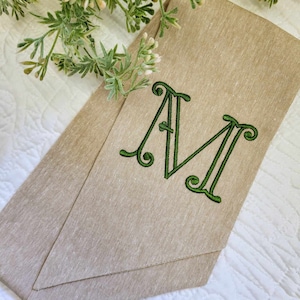 May include: A tan linen-textured fabric strip featuring a large, embroidered letter M in a dark green thread. The letter has a decorative, swirling serif font style. The fabric is folded into a pointed shape, showcasing a woven, natural appearance.