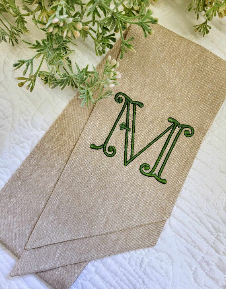 May include: A tan linen-textured fabric strip featuring a large, embroidered letter M in a dark green thread. The letter has a decorative, swirling serif font style. The fabric is folded into a pointed shape, showcasing a woven, natural appearance.