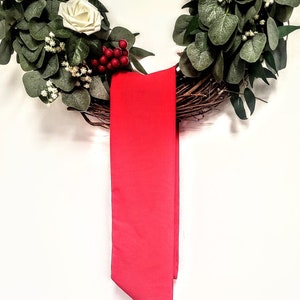 May include: A decorative wreath featuring white roses, green foliage, and red berries. A vibrant red ribbon hangs from the wreath, adding a pop of color. The wreath is made of natural twigs and is perfect for seasonal home decor.