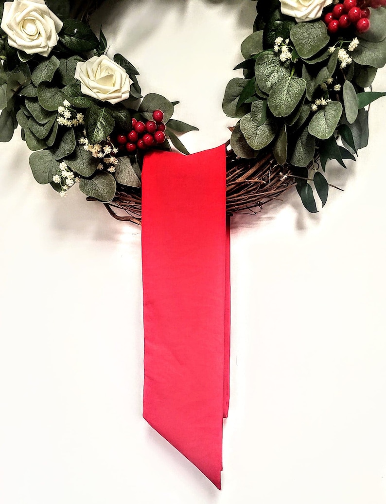 May include: A decorative wreath featuring white roses, green foliage, and red berries. A vibrant red ribbon hangs from the wreath, adding a pop of color. The wreath is made of natural twigs and is perfect for seasonal home decor.