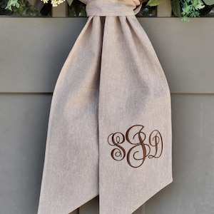 May include: A beige fabric door hanger featuring a knotted top and two hanging tails. The right tail displays a dark brown embroidered monogram with the letters S, S, and D in a decorative script font, centered on the lower portion of the fabric.