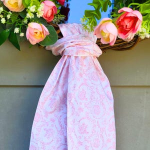 May include: A decorative wreath featuring artificial pink and yellow flowers with green foliage, accented by a long, knotted fabric ribbon in a light pink color with a delicate white floral pattern that hangs down from the center of the wreath.