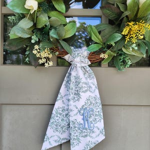 May include: A round wreath featuring green leaves, white tulip-like flowers, and small yellow floral clusters. A white fabric ribbon with a green botanical pattern and a blue embroidered letter H is tied in a knot at the bottom of the wreath.