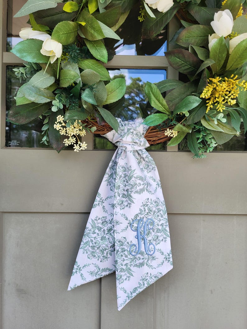 May include: A round wreath featuring green leaves, white tulip-like flowers, and small yellow floral clusters. A white fabric ribbon with a green botanical pattern and a blue embroidered letter H is tied in a knot at the bottom of the wreath.