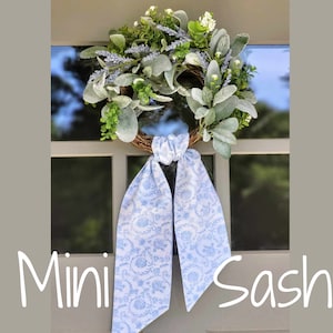 May include: A light blue mini sash with a floral pattern, tied around a green and white wreath with white flowers. The wreath is hanging on a door.