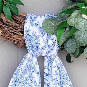 May include: A decorative wreath constructed from brown intertwined twigs and green foliage, featuring a white fabric ribbon with a blue floral toile pattern tied around the center. The ribbon hangs down in two long tails from the wreath base.