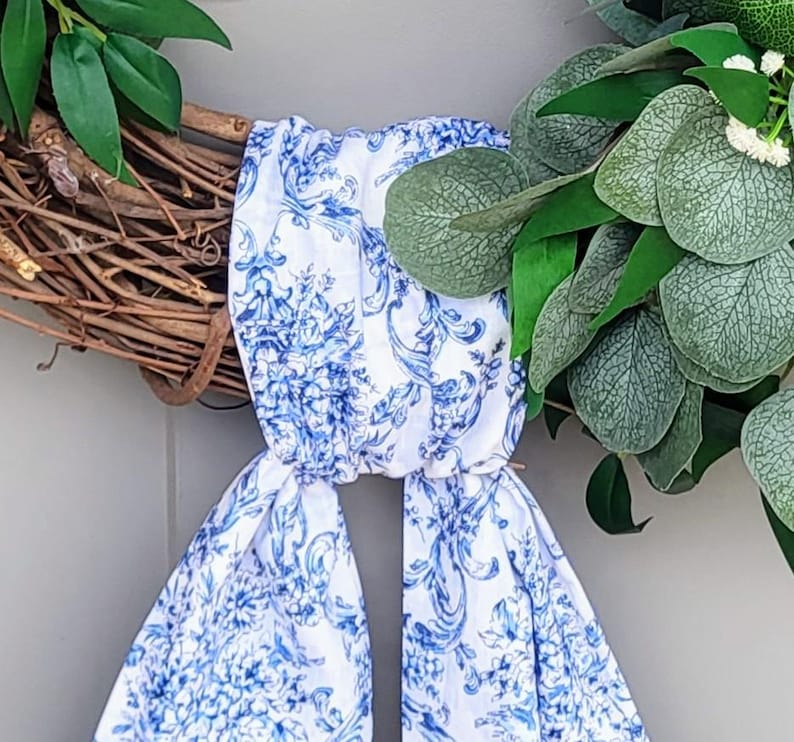 May include: A decorative wreath constructed from brown intertwined twigs and green foliage, featuring a white fabric ribbon with a blue floral toile pattern tied around the center. The ribbon hangs down in two long tails from the wreath base.