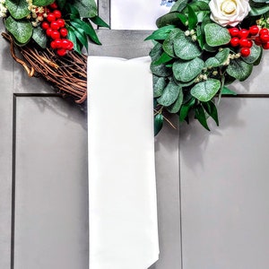 May include: A decorative wreath with white roses, red berries, and green foliage. The wreath is made of woven branches and is adorned with a long, white satin ribbon. The wreath is hanging on a gray door.