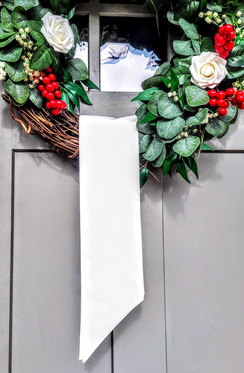 May include: A decorative wreath with white roses, red berries, and green foliage. The wreath is made of woven branches and is adorned with a long, white satin ribbon. The wreath is hanging on a gray door.