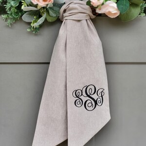 May include: A beige fabric ribbon tied in a knot, featuring a black embroidered monogram with the letters S, S, and J in a decorative script font. The ribbon is positioned below a wreath adorned with light pink roses and green eucalyptus leaves.