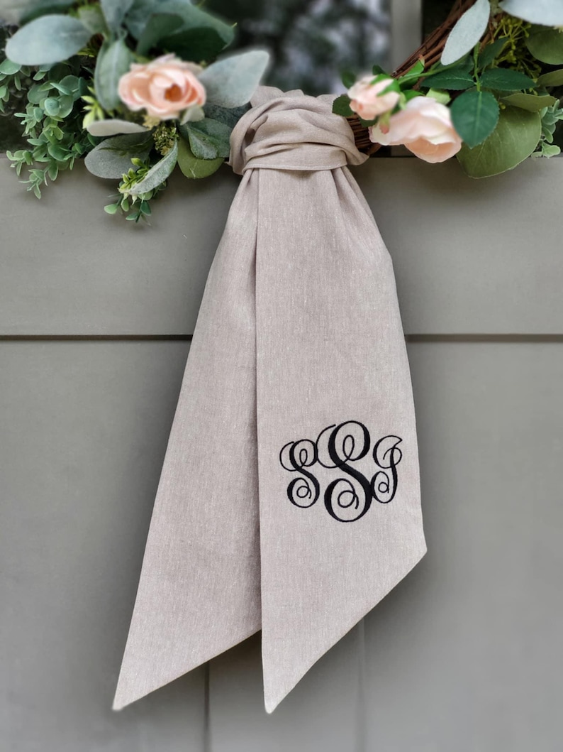 May include: A beige fabric ribbon tied in a knot, featuring a black embroidered monogram with the letters S, S, and J in a decorative script font. The ribbon is positioned below a wreath adorned with light pink roses and green eucalyptus leaves.