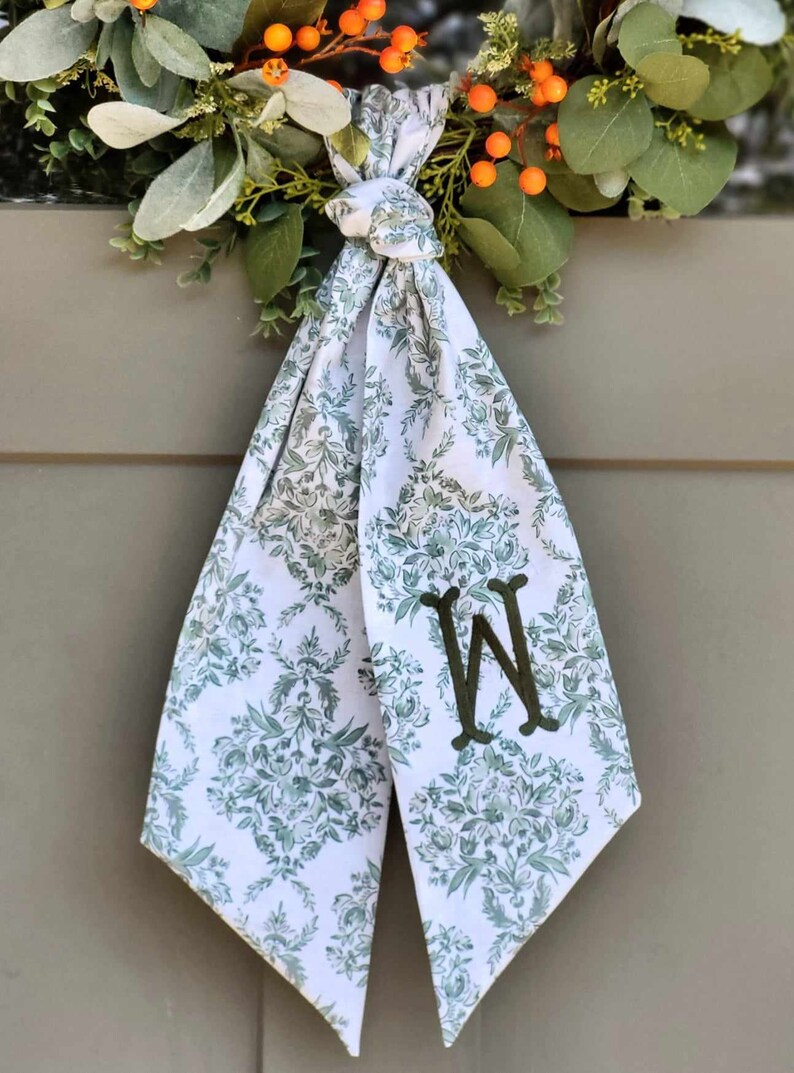 Pu&ograve; includere: Una decorazione in tessuto bianco da appendere con un motivo floreale damascato verde e una lettera W ricamata in verde scuro. Il tessuto &egrave; annodato in alto e scende in due punte, posizionato sotto una ghirlanda con foglie verdi e bacche arancioni.