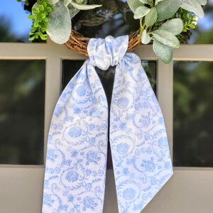 May include: A decorative wreath featuring a brown twig base adorned with green and silver-toned foliage. A long, light blue and white fabric ribbon with a repeating floral and vine pattern is tied in a knot at the center of the wreath.