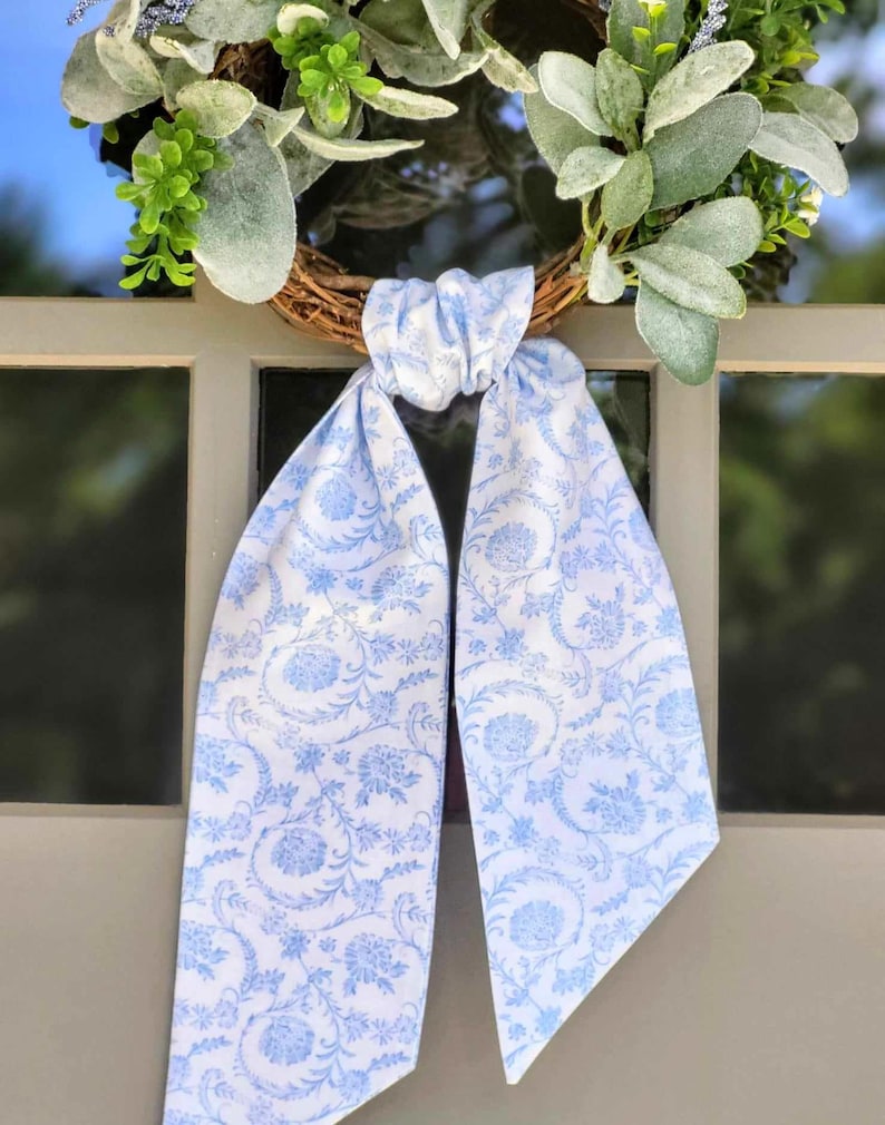 May include: A decorative wreath featuring a brown twig base adorned with green and silver-toned foliage. A long, light blue and white fabric ribbon with a repeating floral and vine pattern is tied in a knot at the center of the wreath.