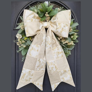 May include: A decorative wreath featuring a large, fabric bow with a patchwork design in cream and gold tones. The bow is layered over a circular base of green foliage, small white berries, and light-colored floral accents, creating a layered look.