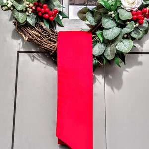 May include: A decorative wreath with white roses, red berries, and green foliage. A bright red ribbon hangs vertically from the wreath, adding a pop of color. The wreath is displayed on a gray door, creating a festive holiday decoration.