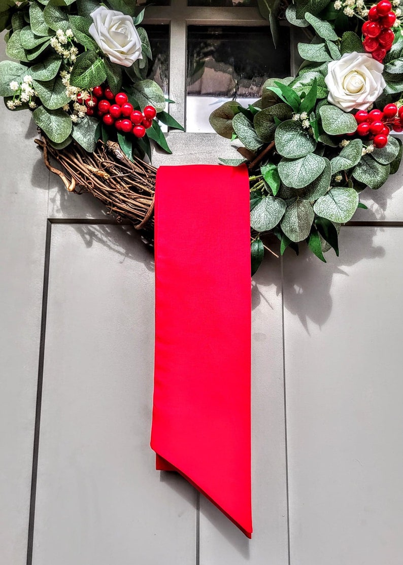 May include: A decorative wreath with white roses, red berries, and green foliage. A bright red ribbon hangs vertically from the wreath, adding a pop of color. The wreath is displayed on a gray door, creating a festive holiday decoration.