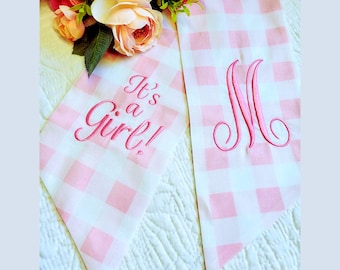 May include: Two pink and white checkered fabric banners. One banner has the embroidered phrase "It's a Girl!" in pink script. The other banner has a pink embroidered letter "M". A bouquet of pink and peach flowers is in the background.