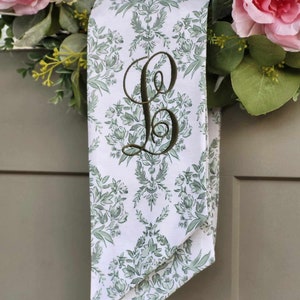 Pu&ograve; includere: Un nastro di tessuto bianco con un motivo floreale damascato verde e una grande lettera B ricamata in verde scuro in corsivo. Il nastro &egrave; drappeggiato su una ghirlanda decorata con rose rosa e fogliame verde su una porta grigia.