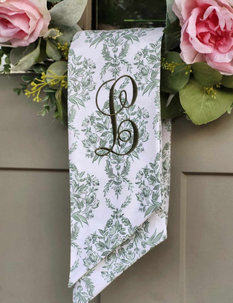 Pu&ograve; includere: Un nastro di tessuto bianco con un motivo floreale damascato verde e una grande lettera B ricamata in verde scuro in corsivo. Il nastro &egrave; drappeggiato su una ghirlanda decorata con rose rosa e fogliame verde su una porta grigia.