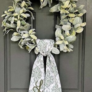 Pu&ograve; includere: Una ghirlanda circolare composta da foglie di orecchia d'agnello verde salvia. Un lungo nastro di tessuto bianco con un motivo damascato floreale grigio ripetuto pende dal basso. La lettera L &egrave; ricamata con filo verde scuro sul nastro.