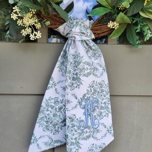 May include: A decorative wreath featuring green foliage and small yellow flowers, accented with a long, white fabric ribbon tied in a knot. The ribbon displays a green floral pattern and features a large, embroidered light blue letter K monogram.