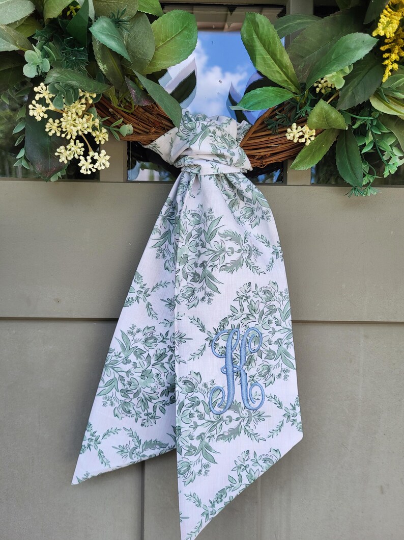 May include: A decorative wreath featuring green foliage and small yellow flowers, accented with a long, white fabric ribbon tied in a knot. The ribbon displays a green floral pattern and features a large, embroidered light blue letter K monogram.