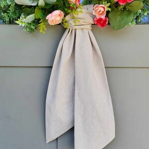 May include: A floral wreath featuring pink and peach-colored artificial roses, green foliage, and silver-green lamb's ear leaves. A long, solid light beige fabric ribbon is tied in a knot at the center, with two long tails hanging downward.