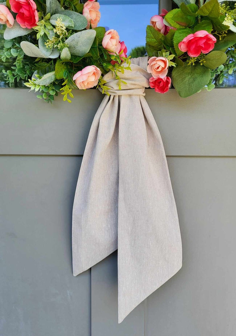 May include: A floral wreath featuring pink and peach-colored artificial roses, green foliage, and silver-green lamb's ear leaves. A long, solid light beige fabric ribbon is tied in a knot at the center, with two long tails hanging downward.