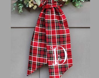 May include: A red, white, and black plaid fabric ribbon tied in a knot, featuring a large white embroidered letter D in a decorative script font. The ribbon is attached to a green leafy wreath with small white flowers at the top.