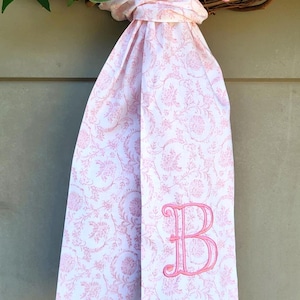 May include: A white fabric ribbon with a pink floral pattern and a pink embroidered letter B monogram. The ribbon is tied in a knot and hangs from a circular wreath decorated with pink and peach-colored artificial flowers and green leaves.
