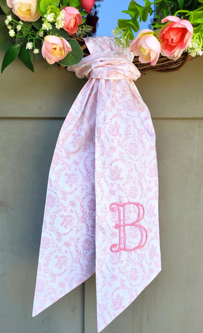 May include: A white fabric ribbon with a pink floral pattern and a pink embroidered letter B monogram. The ribbon is tied in a knot and hangs from a circular wreath decorated with pink and peach-colored artificial flowers and green leaves.