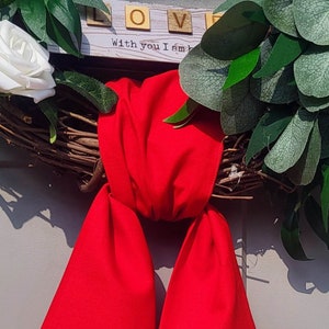 May include: A grapevine wreath adorned with white roses, green foliage, and a vibrant red bow. The word "LOVE" is spelled out with wooden letter tiles, and the phrase "With you I am" is printed below.