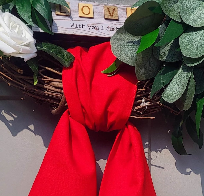 May include: A grapevine wreath adorned with white roses, green foliage, and a vibrant red bow. The word "LOVE" is spelled out with wooden letter tiles, and the phrase "With you I am" is printed below.