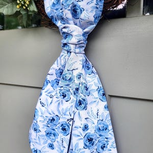 May include: A long fabric ribbon featuring a white background with a repeating pattern of blue roses and light blue leaves. The ribbon is tied in a knot at the top and hangs downward, displaying a floral print with various shades of blue and white.