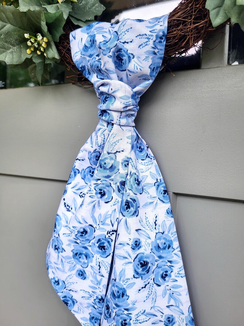 May include: A long fabric ribbon featuring a white background with a repeating pattern of blue roses and light blue leaves. The ribbon is tied in a knot at the top and hangs downward, displaying a floral print with various shades of blue and white.