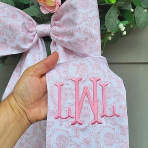 May include: A large decorative bow made of white fabric with a light pink floral pattern. The fabric features a prominent pink embroidered monogram with the letters LWL. The bow is positioned in front of a wreath with pink roses and green foliage.