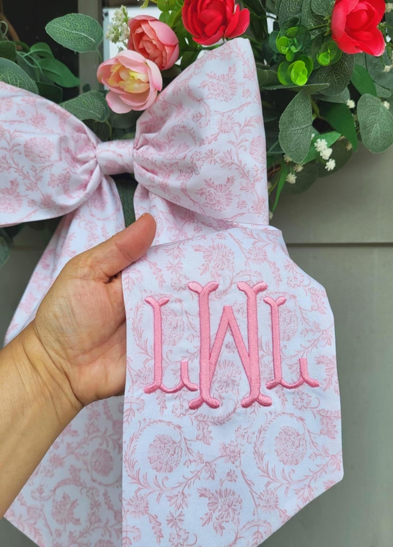 May include: A large decorative bow made of white fabric with a light pink floral pattern. The fabric features a prominent pink embroidered monogram with the letters LWL. The bow is positioned in front of a wreath with pink roses and green foliage.