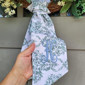 May include: A white fabric ribbon featuring a green botanical floral pattern and a light blue embroidered letter K monogram. The ribbon is tied in a knot and hangs down, displaying the intricate stitching of the letter and the floral print.