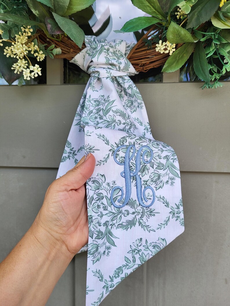 May include: A white fabric ribbon featuring a green botanical floral pattern and a light blue embroidered letter K monogram. The ribbon is tied in a knot and hangs down, displaying the intricate stitching of the letter and the floral print.