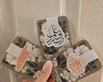 May include: Three decorative bookmarks with floral designs and Arabic calligraphy. The bookmarks are in clear plastic packaging. One bookmark has a pink book sticker with text. The other two have white bands with Arabic calligraphy.