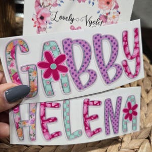 May include: Two rectangular vinyl stickers featuring the names GABBY and HELENA in large, colorful, patterned block letters. The letters incorporate floral, polka dot, and checkered designs in shades of pink, purple, and teal, with a pink flower icon.