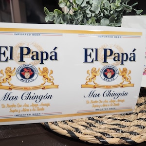 May include: White beer label with the text "El Papá" in navy blue, featuring a gold emblem and the words "Mas Chingón." The label also includes the text "Imported Beer" and "Cerveceria Modelo Mexico."