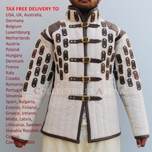 Puede incluir: Una chaqueta acolchada de color crema con detalles de cuero marrón y hebillas de latón. La chaqueta tiene un cuello alto y está adornada con numerosos parches de cuero. El texto "COLLECTIBLES ARENA" es visible en la chaqueta.