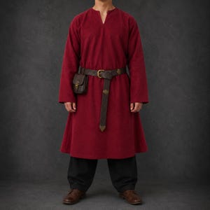 以下が含まれることがあります： Vネックの深い赤色の長袖膝丈チュニックに、黒いズボンと茶色の革靴を合わせています。真鍮のバックルが付いたダークブラウンの型押しレザーベルトと、お揃いの小さなレザーポーチを腰に巻いています。