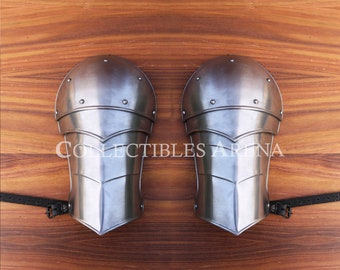 May include: A pair of silver metal elbow guards with a textured surface and a strap on each side. The guards are on a wooden surface.