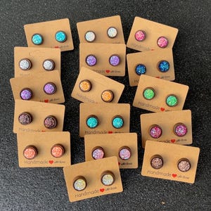 May include: A collection of glitter stud earrings in various colors, including blue, silver, purple, orange, green, pink, and brown. Each pair is mounted on a small brown card with the text "Handmade with love".