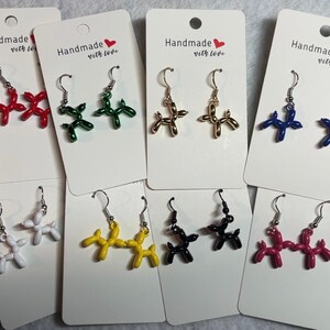 May include: A collection of handmade balloon dog earrings in various colors, including red, green, gold, blue, white, yellow, black, and pink. Each pair is displayed on a white card with the text "Handmade with love".