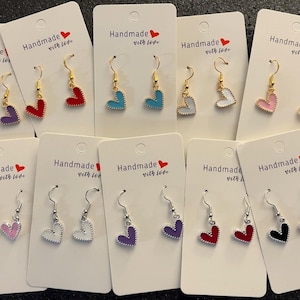 May include: A collection of heart-shaped earrings in various colors, including purple, red, blue, white, pink, and black. The earrings are displayed on white cards with the text "Handmade with love". The earrings have gold or silver hooks.